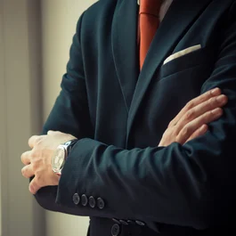 Uomo in giacca blu e cravatta arancione con braccia incrociate vicino alla finestra